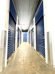 A well-lit hallway of a storage facility with blue roll-up doors on either side, white walls, and a high ceiling. The polished concrete floor reflects the light, creating a clean and organized appearance.